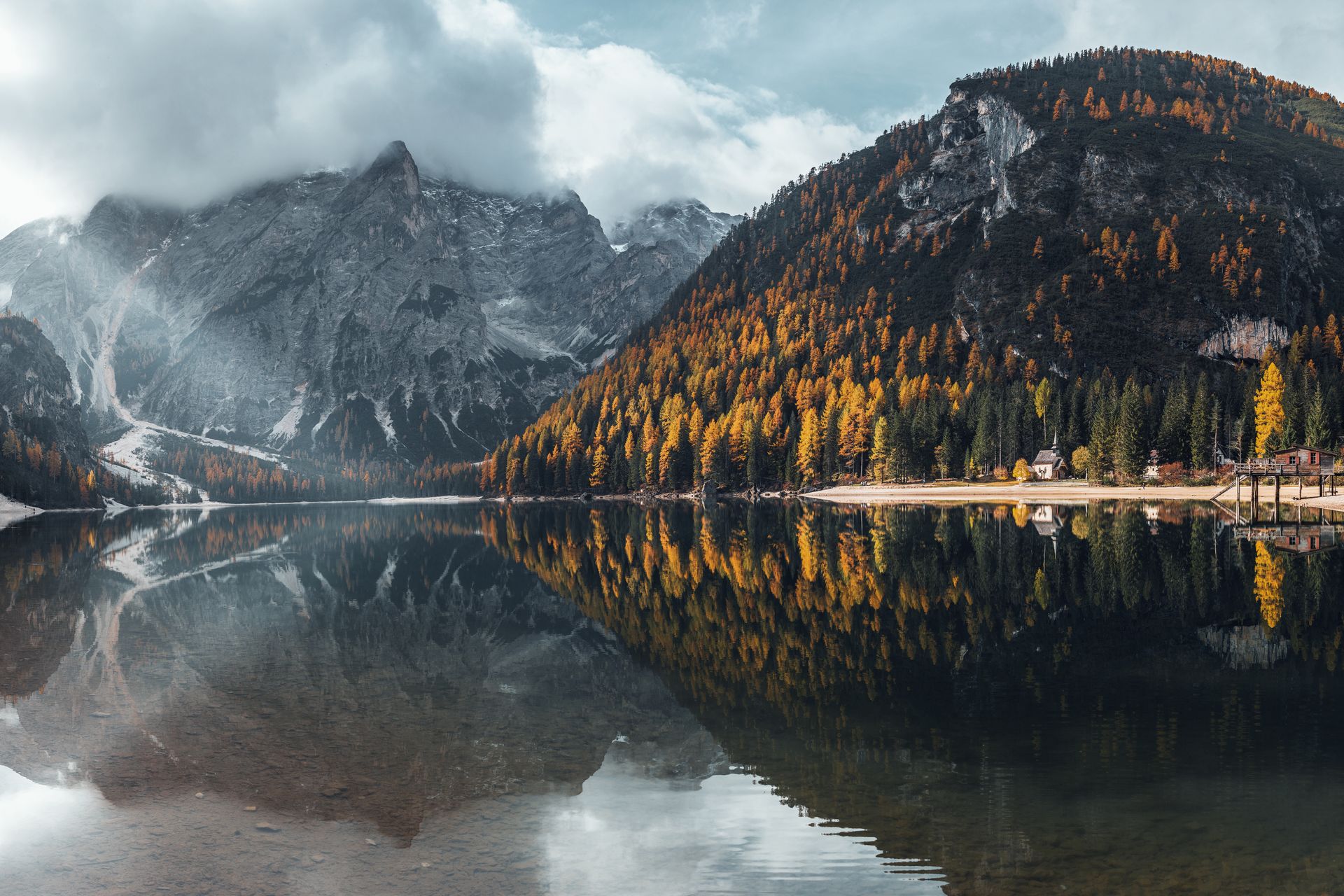 Lago di Braies in autunno con larici dorati, montagne avvolte dalle nuvole e riflessi nell'acqua