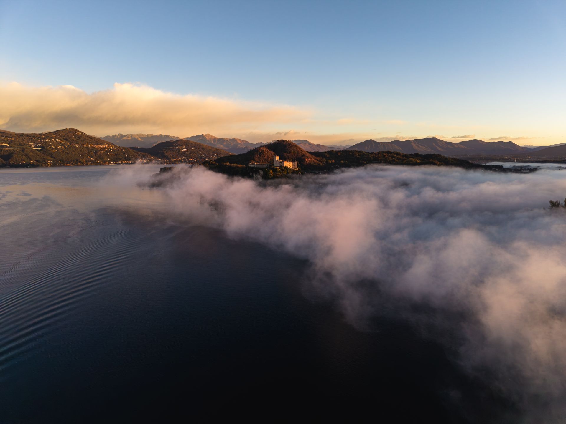 Lago d'Orta avvolto dalla nebbia all'alba, con colline e montagne illuminate dalla luce del mattino