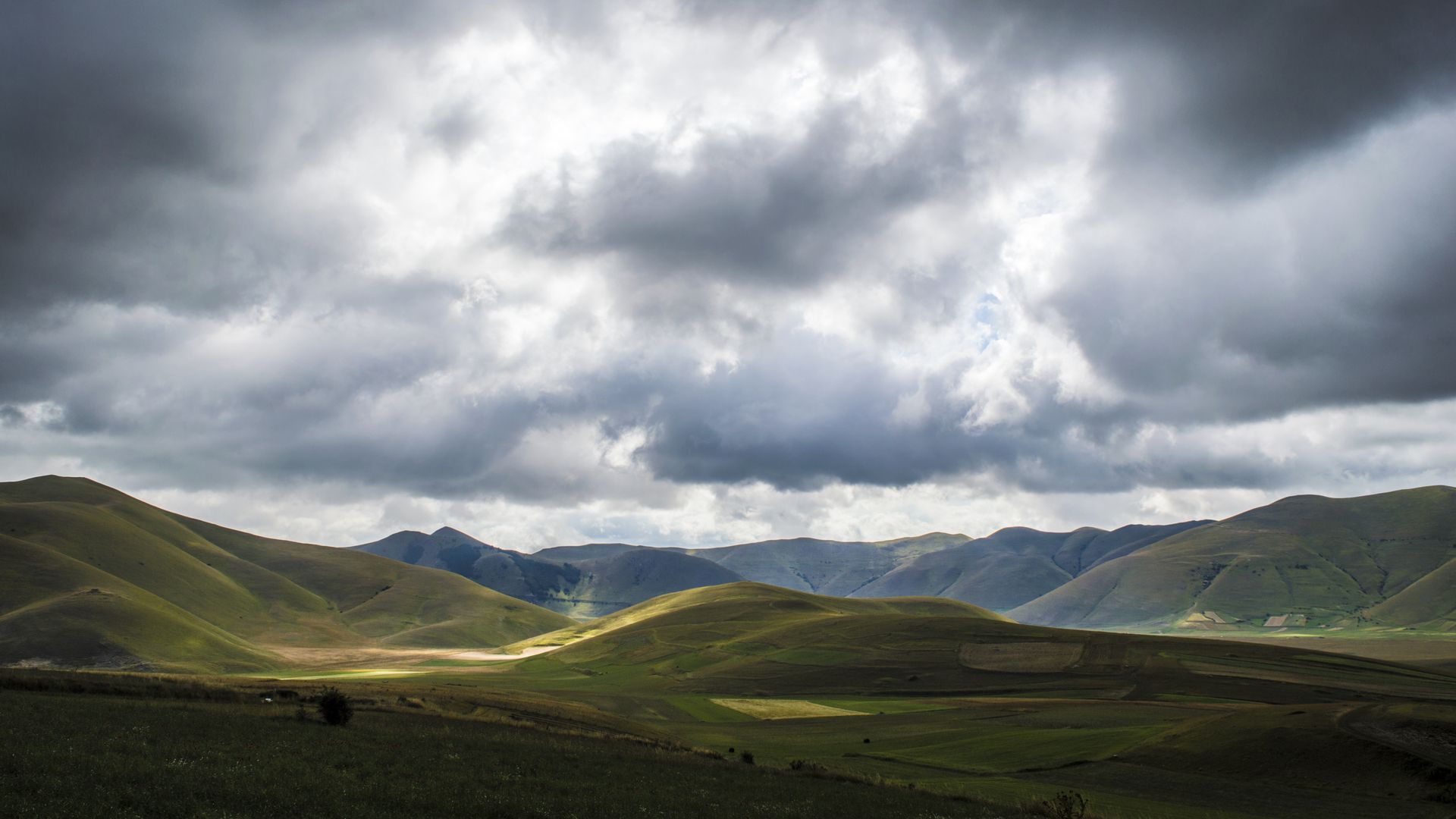 Colline ondulate del Pian Grande di Castelluccio con nuvole grigie e giochi di luce sul paesaggio